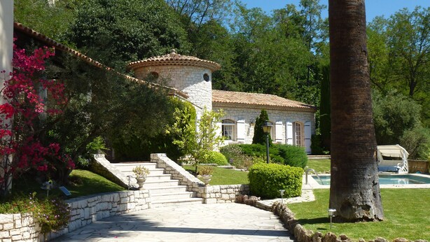 Uma escadaria de pedra leva a uma casa com telhado de telhas, cercada por vegetação exuberante e uma piscina.