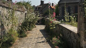 Ein schmaler Kopfsteinpflasterweg, gesäumt von Steinmauern und blühenden Pflanzen, führt zu traditionellen Steinhäusern mit Satteldächern.