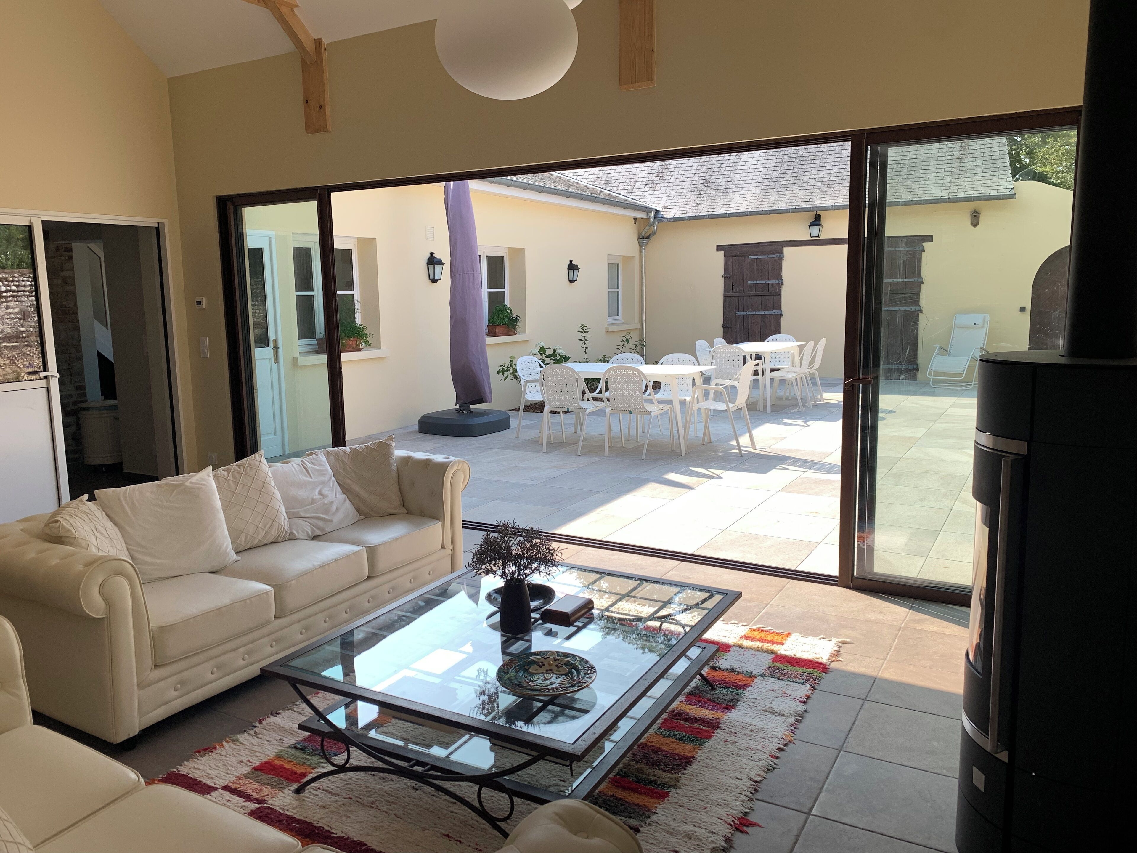 A modern living room with a glass coffee table, white sofa, and a view of an outdoor patio with white chairs and a barbecue grill.