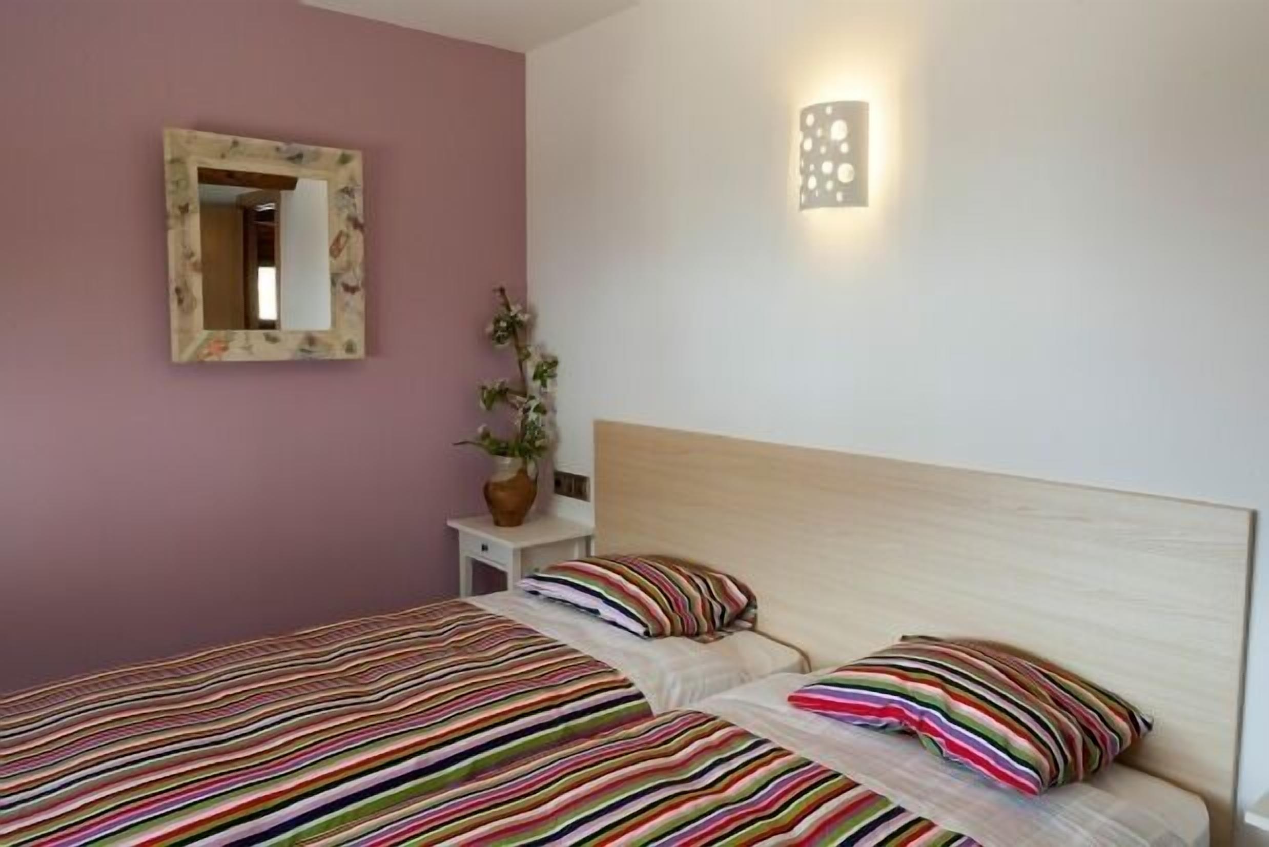 A bedroom with two beds, a mirror, a wall-mounted light fixture, and a small bedside table with a vase of flowers.