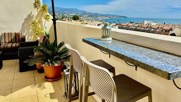 Une terrasse dotée de mobilier d’extérieur, d’un pot de fleurs et offrant une vue sur la mer et le paysage urbain.