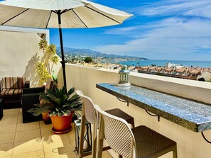 Eine Terrasse mit Gartenmöbeln, einem Blumentopf und Blick auf das Meer und die Stadt.