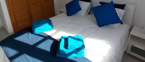 Un lit bien fait, avec des oreillers bleus et une couverture bleue, une armoire en bois et une table de chevet blanche avec une lampe.