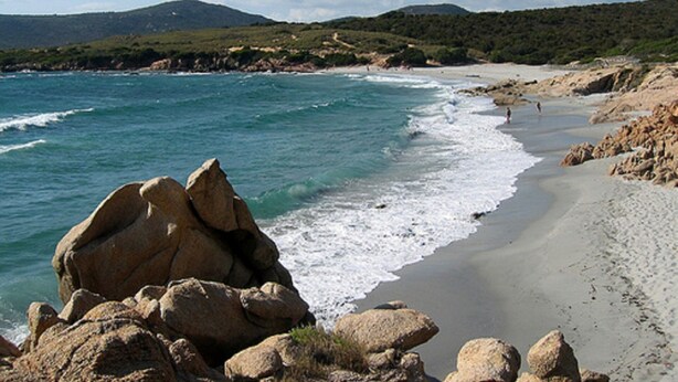A beach with large rocks in the foreground, a sandy shore, and a hilly landscape in the background.