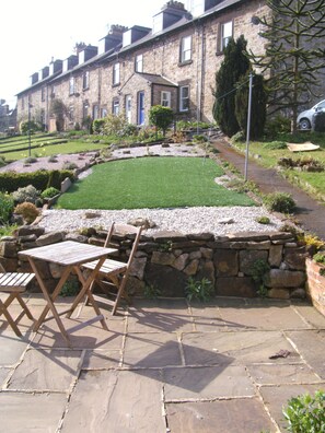 A stone patio with a lawn and a stone wall. There are two wooden chairs and a table.