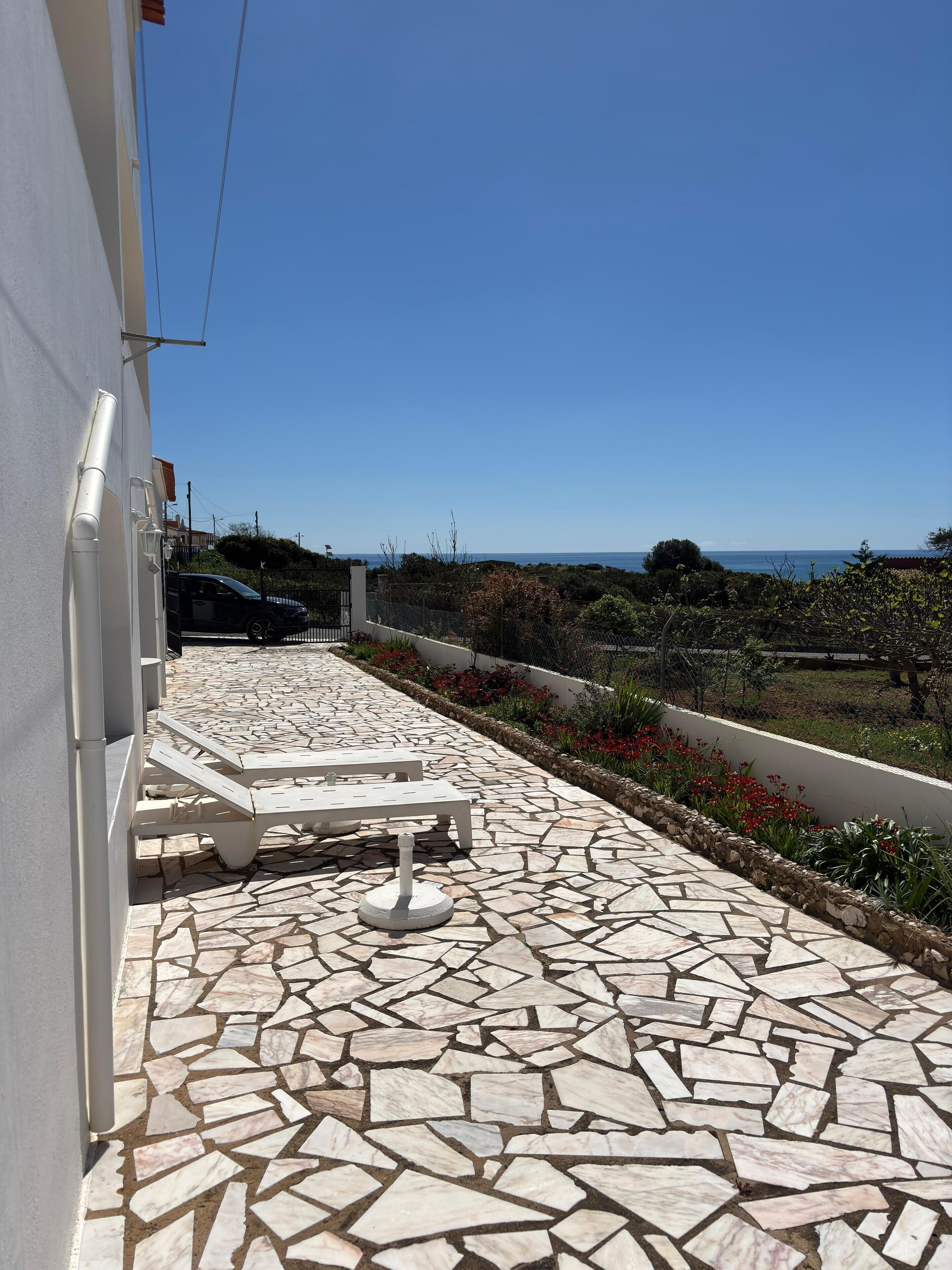 A patio with stone tiles, white lounge chairs, and a view of the sea.