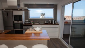 A modern kitchen with a wooden island, white bar stools, and a view of a coastal town through a large window.