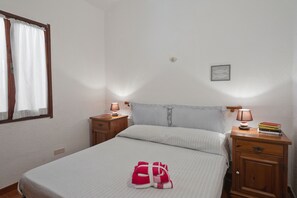 Een net opgemaakt bed met een gestreept dekbed, twee nachtkastjes met lampen, een klein, ingelijst fotootje aan de muur en een raam met gordijnen.