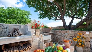 A rustic outdoor setting with a stone wall, a wooden table with fruits and vegetables, a large vase with sunflowers, and a tree in the background. - Puglia Salento luxury villa with swimming pool and hydro spa. a few km from leuca  (alessano)