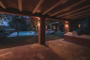 Un patio al aire libre con suelo de piedra, una piscina y una zona de estar cubierta con cojines.