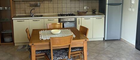 A compact kitchen with a dining table, chairs, a gas stove, a refrigerator, and tiled flooring.