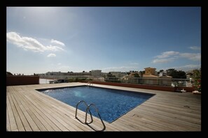 A rooftop swimming pool with a wooden deck, metal pool ladder, and a clear view of the surrounding buildings and sky.