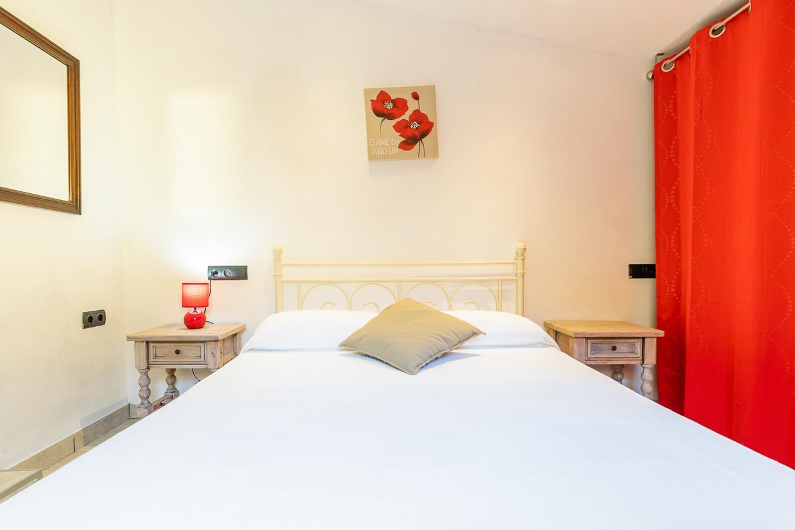 A bedroom with a white bed, a red lamp on the left, a red curtain on the right, and a painting of red flowers on the wall.