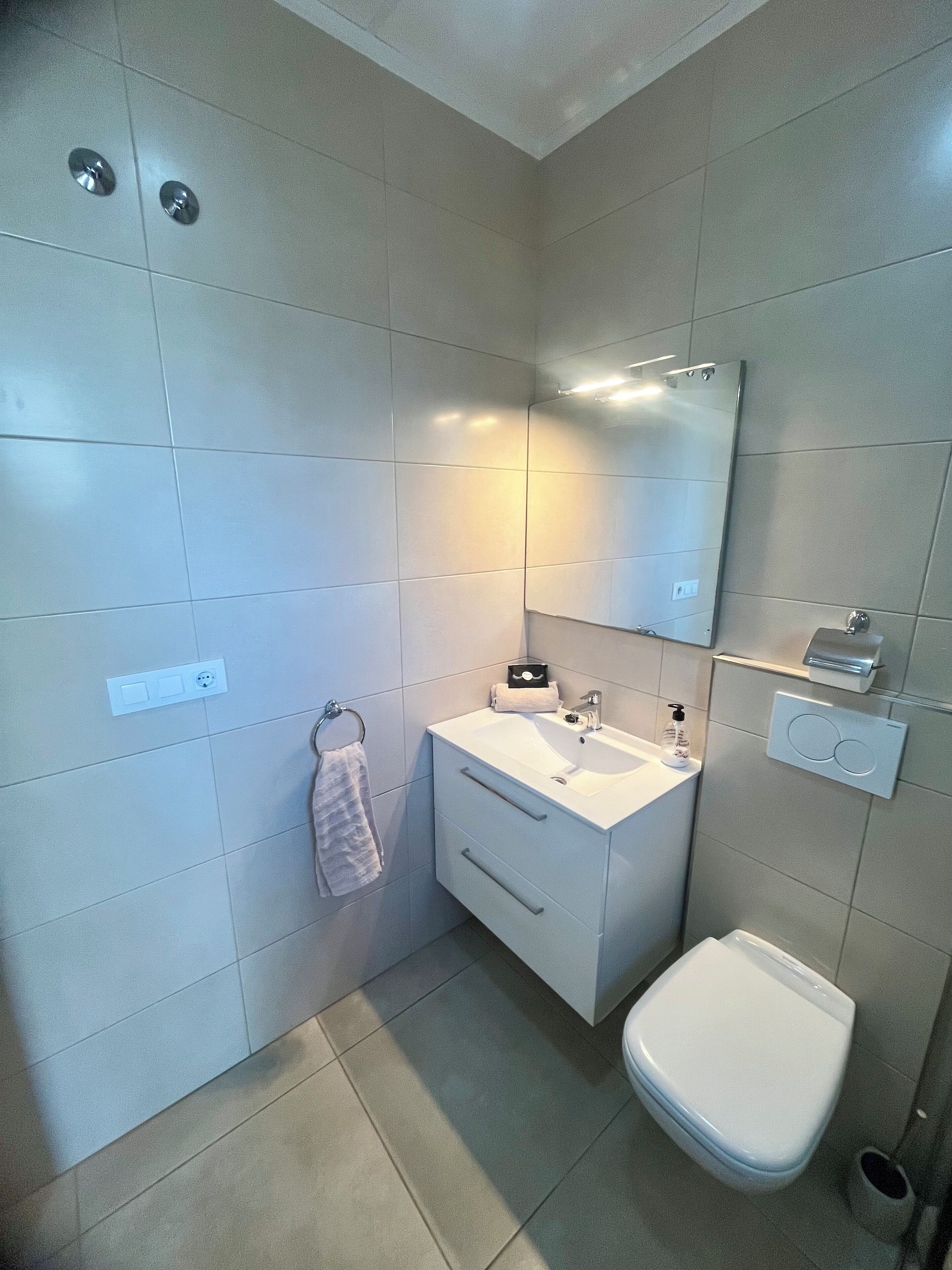 A modern bathroom with a white sink, mirror, and toilet. There is a towel rack with a towel hanging on it, and a soap dispenser on the sink.