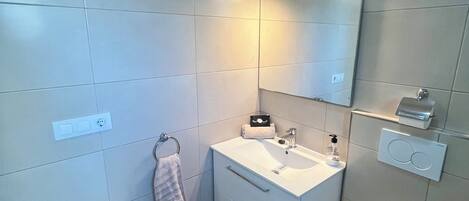 A modern bathroom with a white sink, mirror, and toilet. There is a towel rack with a towel hanging on it, and a soap dispenser on the sink.