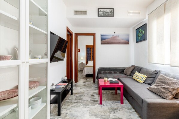 A compact living space with a sofa, a small table, a TV mounted on the wall, and a bookshelf.