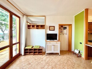 Una habitación con un ventanal grande, un televisor sobre un mueble bajo, un banco con cojines y una pared verde.