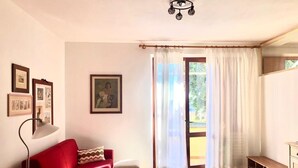 A living room with a red sofa, a small table, and a lamp. There is a window with curtains and a door leading outside.