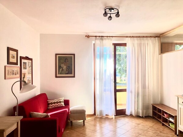Un salón con un sofá rojo, una mesita y una lámpara. Hay una ventana con cortinas y una puerta que da al exterior.
