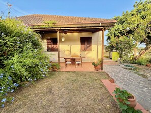 A house with a tiled roof, a patio with a table and chairs, and a garden with potted plants and flowers.