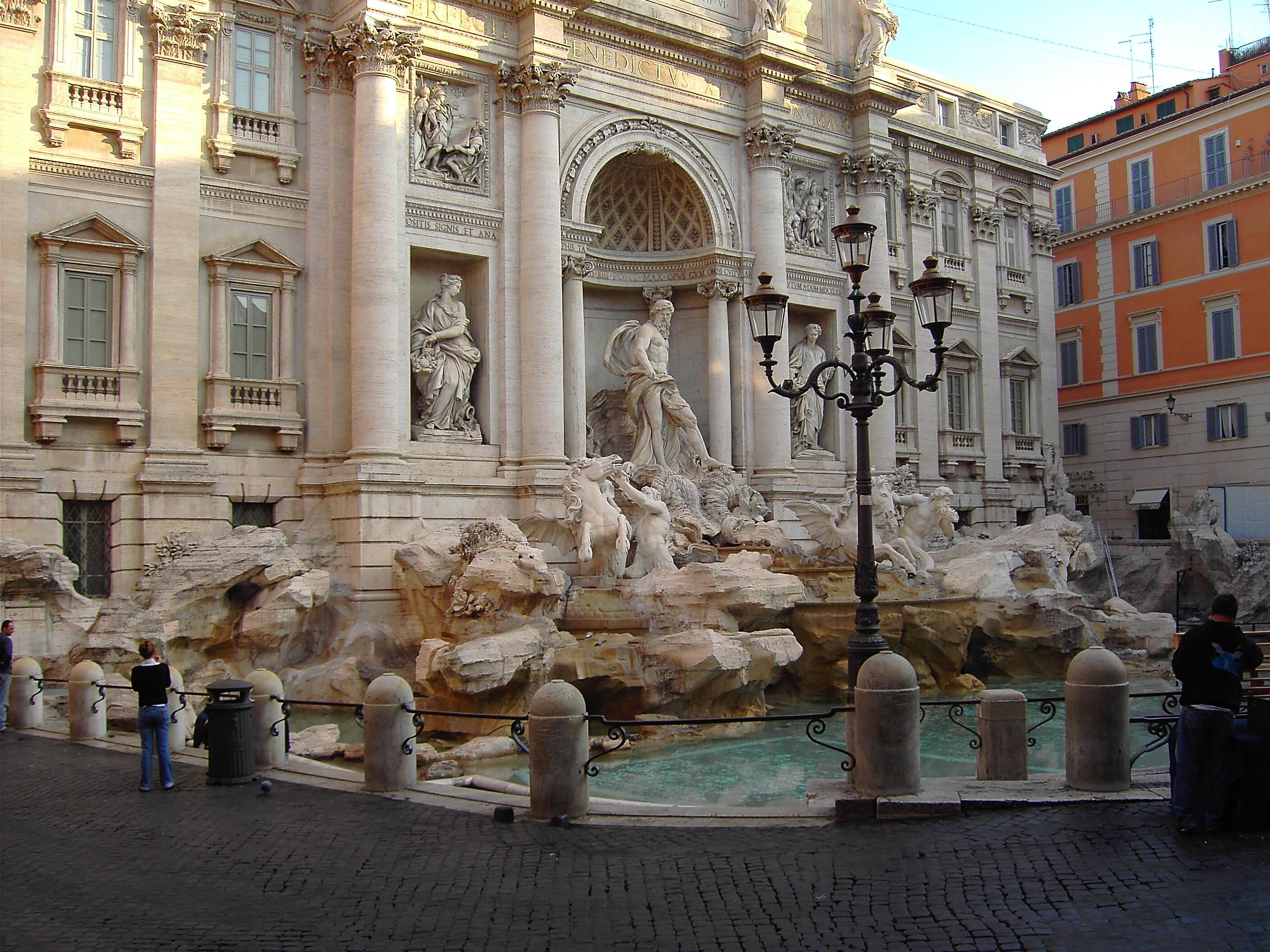 A historic fountain with sculptures, surrounded by classical architecture and a cobblestone street.