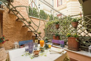 Um pátio externo com mesa posta para refeições, rodeado por plantas em vasos e um guarda-corpo de metal.