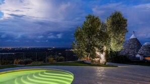 A stone patio with a pool, trees, and a view of a distant cityscape at dusk.