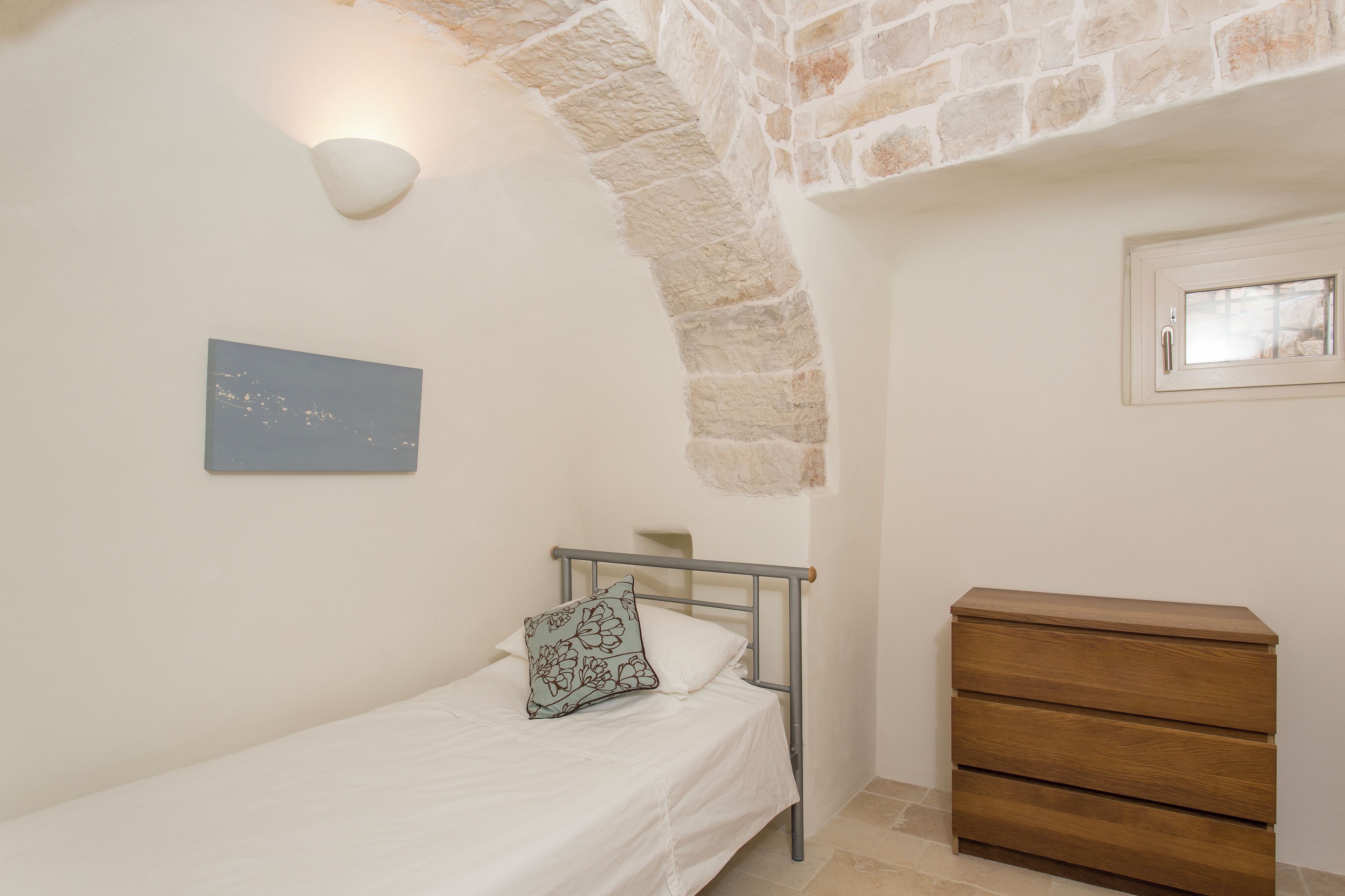 A bedroom with a bed, a dresser, a wall-mounted light, and a window.