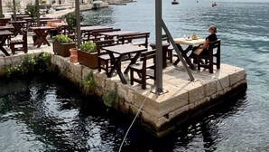 Een restaurant aan het water met terras, een stenen gebouw op de achtergrond en boten op het water.