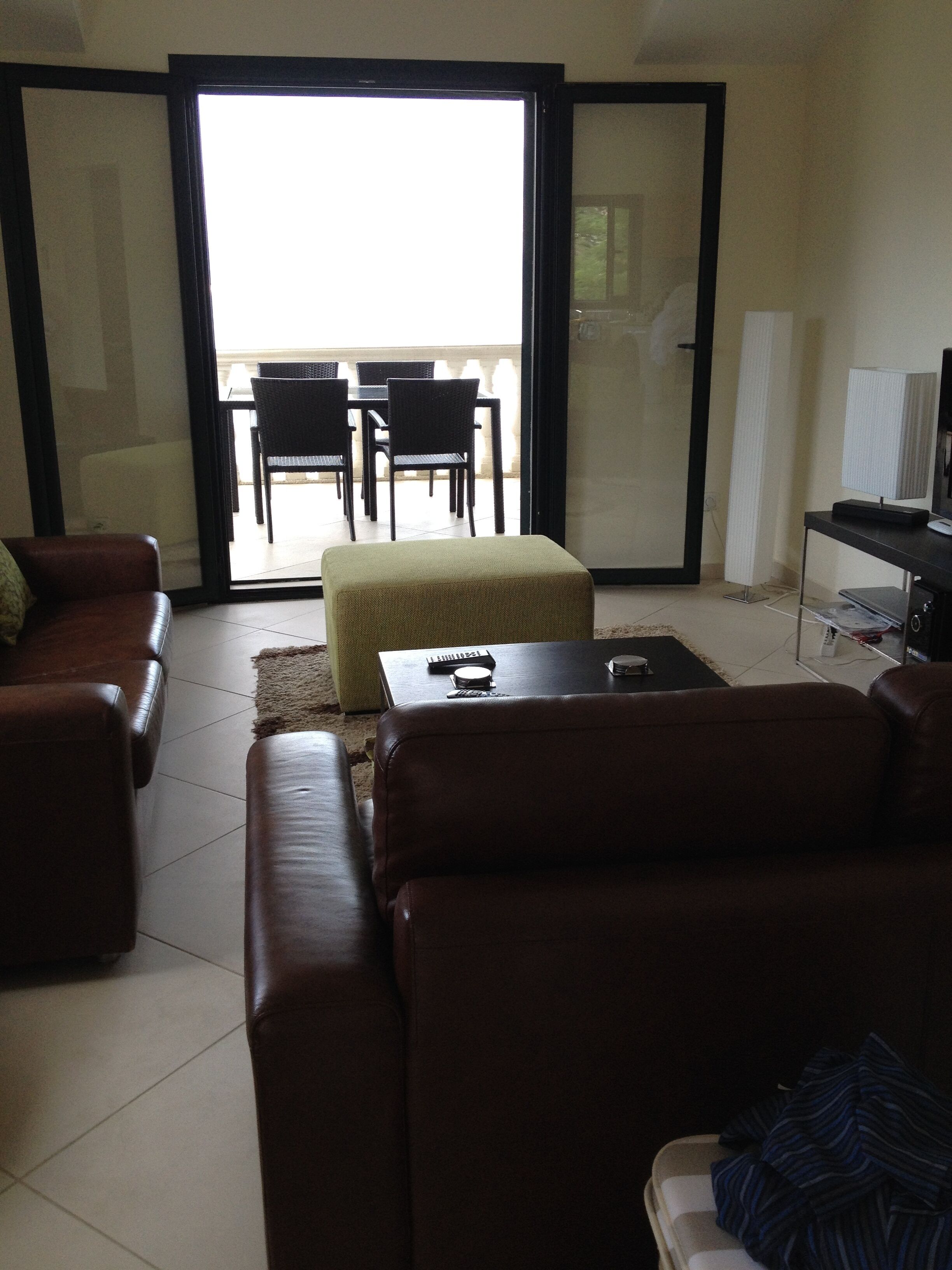 A living room with a brown leather sofa, a small ottoman, a coffee table, and a television on a stand. There is a sliding glass door leading to an outdoor area with a table and chairs.
