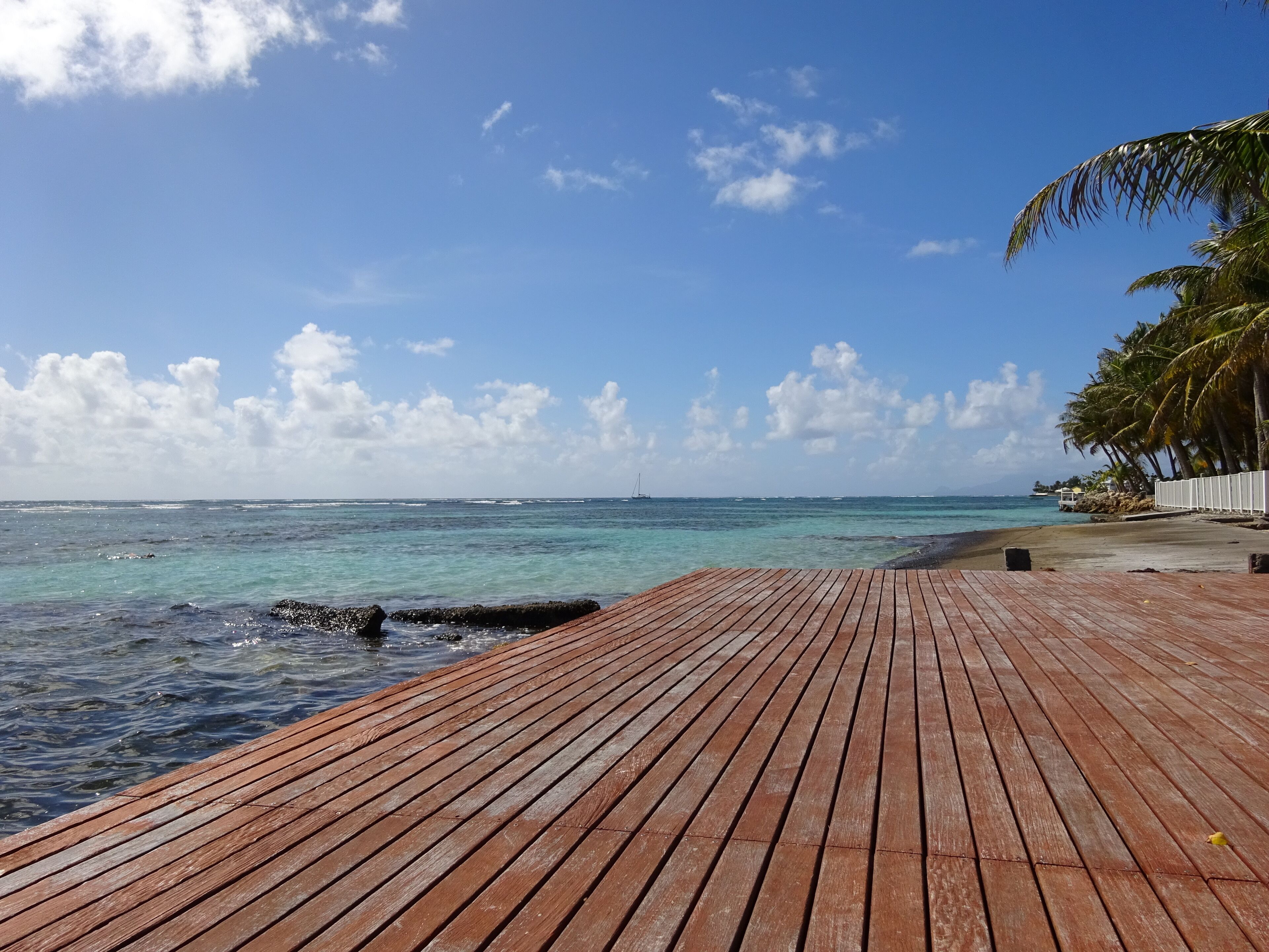 Een houten terras dat naar een strand leidt met helder turquoise water, palmbomen en een zeilboot in de verte.