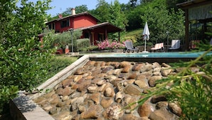 A swimming pool with a stone waterfall feature, surrounded by a garden with various plants and a red house in the background.