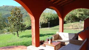 An outdoor patio with wicker furniture, a wooden table, and a view of a green landscape and hills.