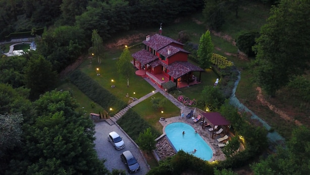 An aerial view of a house with a swimming pool, surrounded by a well-maintained garden and a driveway with parked cars.
