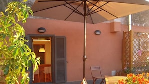 Um pátio externo com mesa, cadeiras, um guarda-sol grande e plantas em vasos.