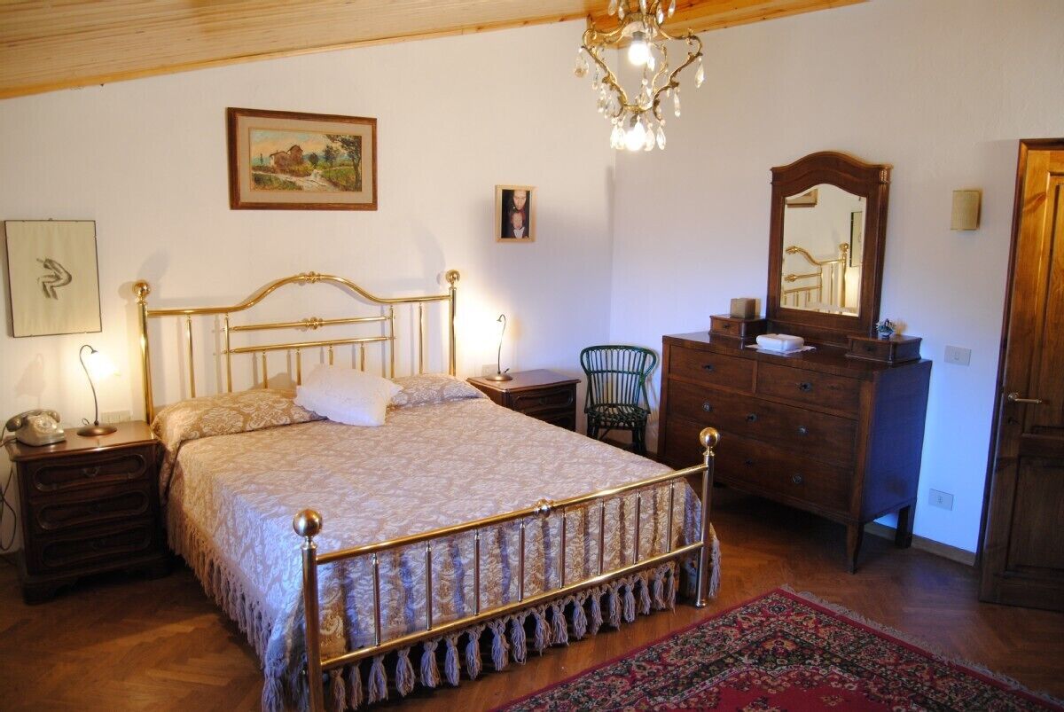 A bedroom with a bed, a dresser, a chair, a nightstand, a lamp, a mirror, and a chandelier.