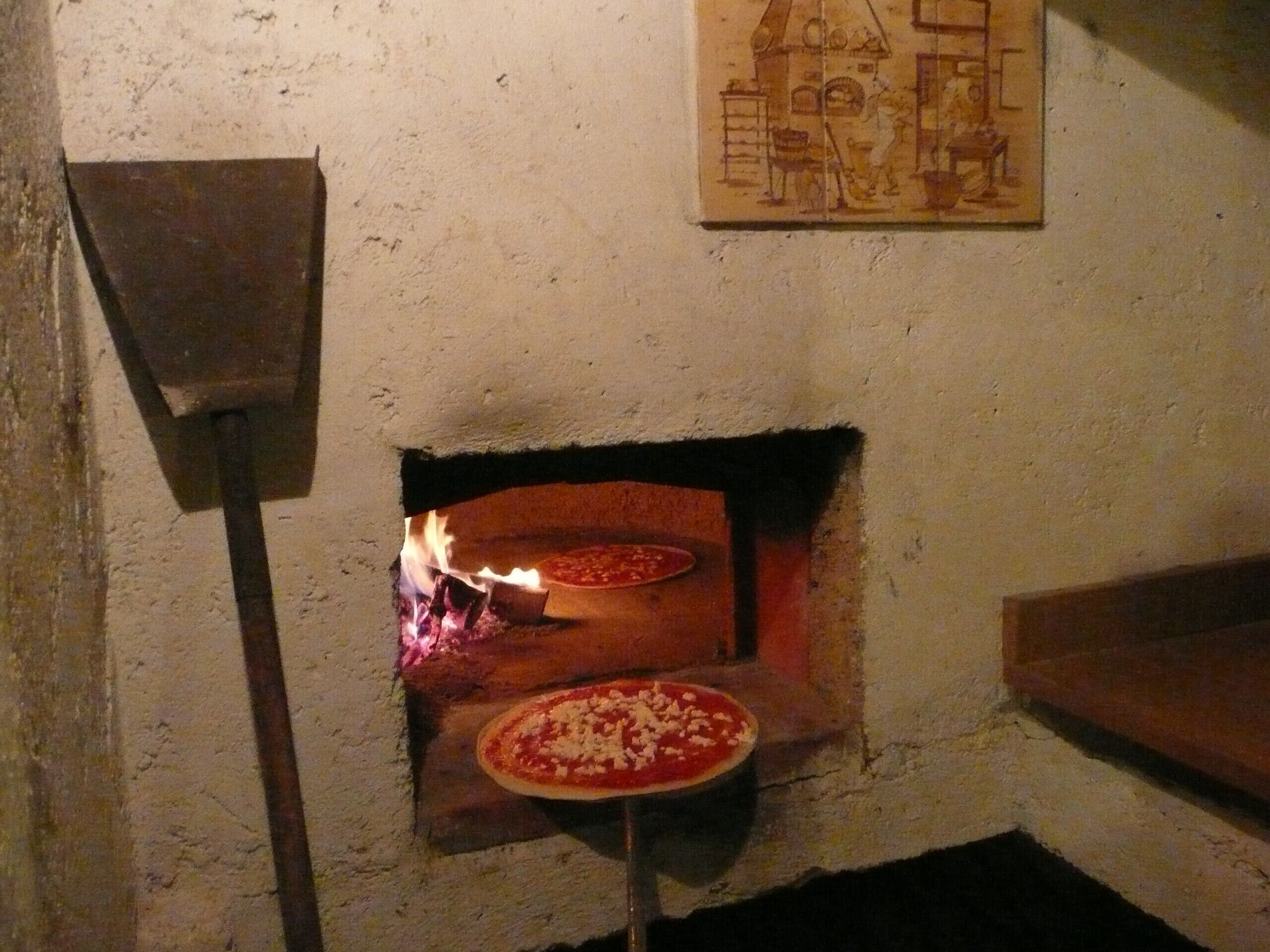 A traditional wood-fired pizza oven with a pizza inside, a pizza peel in front, and a mural on the wall.