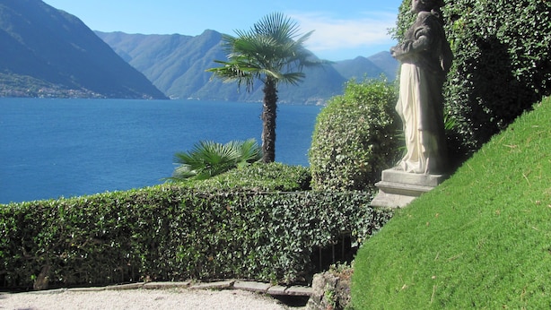 A landscaped area with a statue, a palm tree, and a view of a lake with mountains in the background.