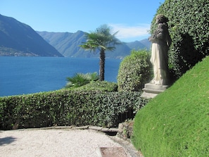 Stroll through the serene lakeside garden, featuring a historic statue and lush greenery, surrounded by a tranquil lake and majestic mountains.