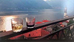 A balcony with two glasses of wine overlooking a lake with mountains and a church in the background.