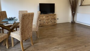 Un soggiorno con tavolo da pranzo, sedie in vimini, un mobile in legno con televisore a schermo piatto e un'alta pianta decorativa.