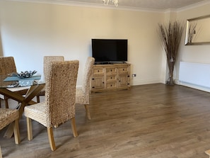 A living room with a dining table, wicker chairs, a wooden cabinet with a flat-screen TV, and a tall decorative plant.