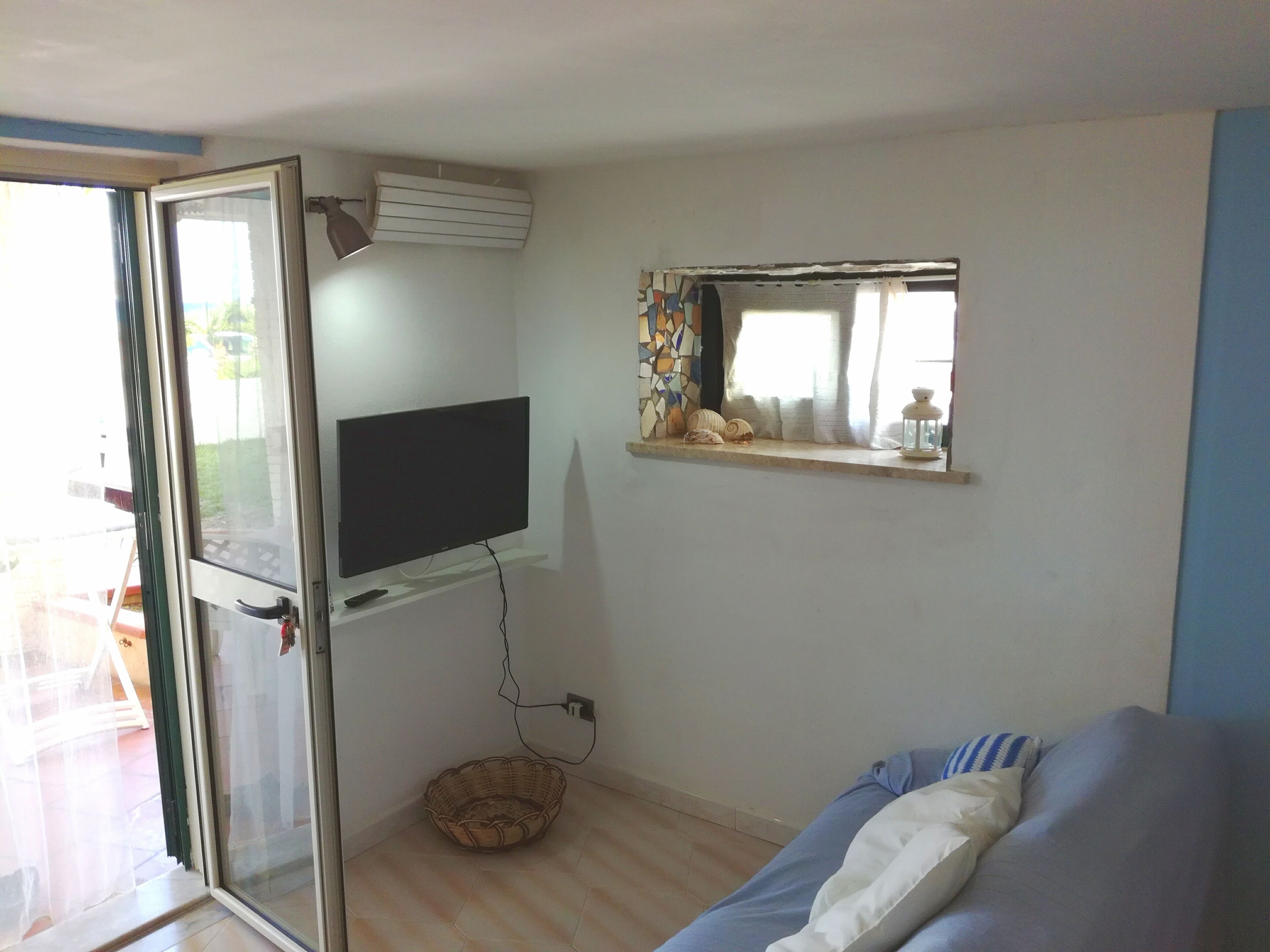 Een kamer met een aan de muur bevestigde televisie, een klein plankje met decoratieve objecten, een rieten mand op de vloer en een bed met een blauw-witte deken.