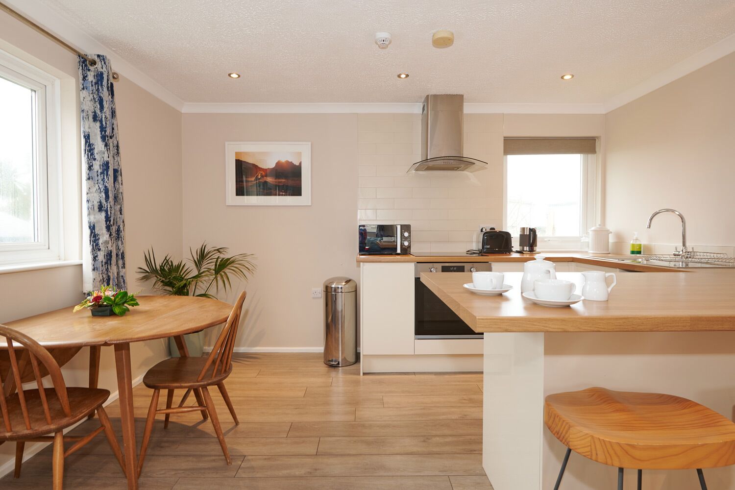 Een moderne keuken met een houten eettafel, stoelen, een klein eilandje met keukengerei, een magnetron, een broodrooster en een spoelbak.