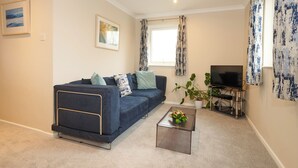 A living room with a dark blue sofa, a coffee table with flowers, a TV on a stand, and a window with curtains. - Spacious 1 bedroomed apartment in the seaside village of Gwithian (Gwithian)