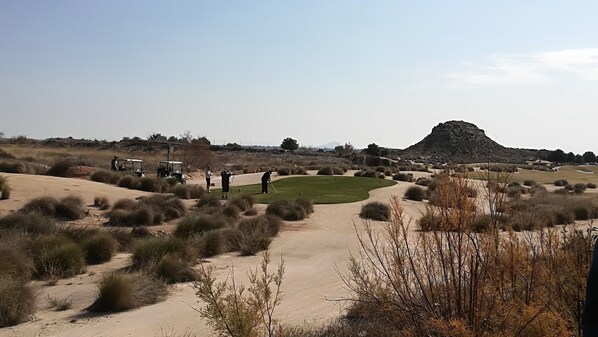 Un parcours de golf situé dans un désert, avec des dunes de sable, une végétation clairsemée et une chaîne de montagnes à l’horizon.