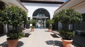 Un patio con árboles en macetas, una entrada central con puertas de vidrio y suelo de baldosas.
