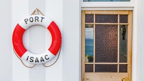 A white wall with a red and white life ring labeled "PORT ISAAC" and a wooden door with a textured glass panel and a sign above it reading "BEACH HOUSE".