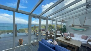 A spacious room with a large glass door leading to a deck with a view of the sea. Inside, there is a blue sofa, a small table, and wicker chairs.