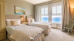 A bedroom with two beds, a painting of a beach scene, a lamp, and a view of the sea through large windows.